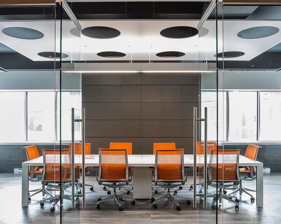 Modern conference room with a glass wall, a long white table, and eight orange office chairs. The room has large windows, gray walls, wood flooring, and a ceiling with round black acoustic panels.
