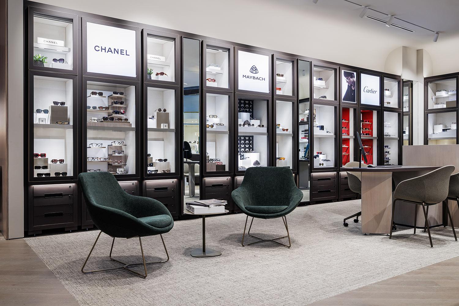 Modern eyewear boutique with glass display cases showcasing designer brands like Chanel, Maybach, and Cartier. Two dark green chairs and a small table sit on a gray rug, creating a stylish seating area.