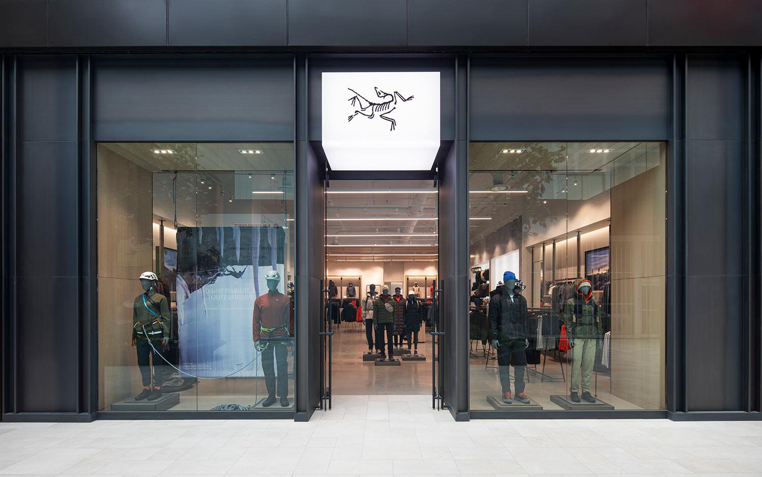 Storefront of an Arc'teryx retail location with large glass windows displaying mannequins in outdoor gear and a white sign above the entrance featuring the brand’s black fossil logo.