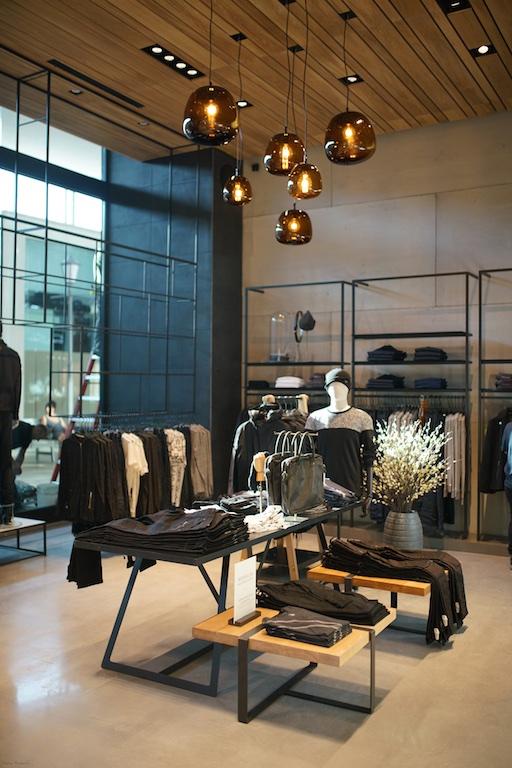 A stylish clothing store interior with hanging pendant lights, black metal racks displaying folded and hanging clothes, mannequins dressed in dark outfits, and a vase of white flowers on a table in the foreground.