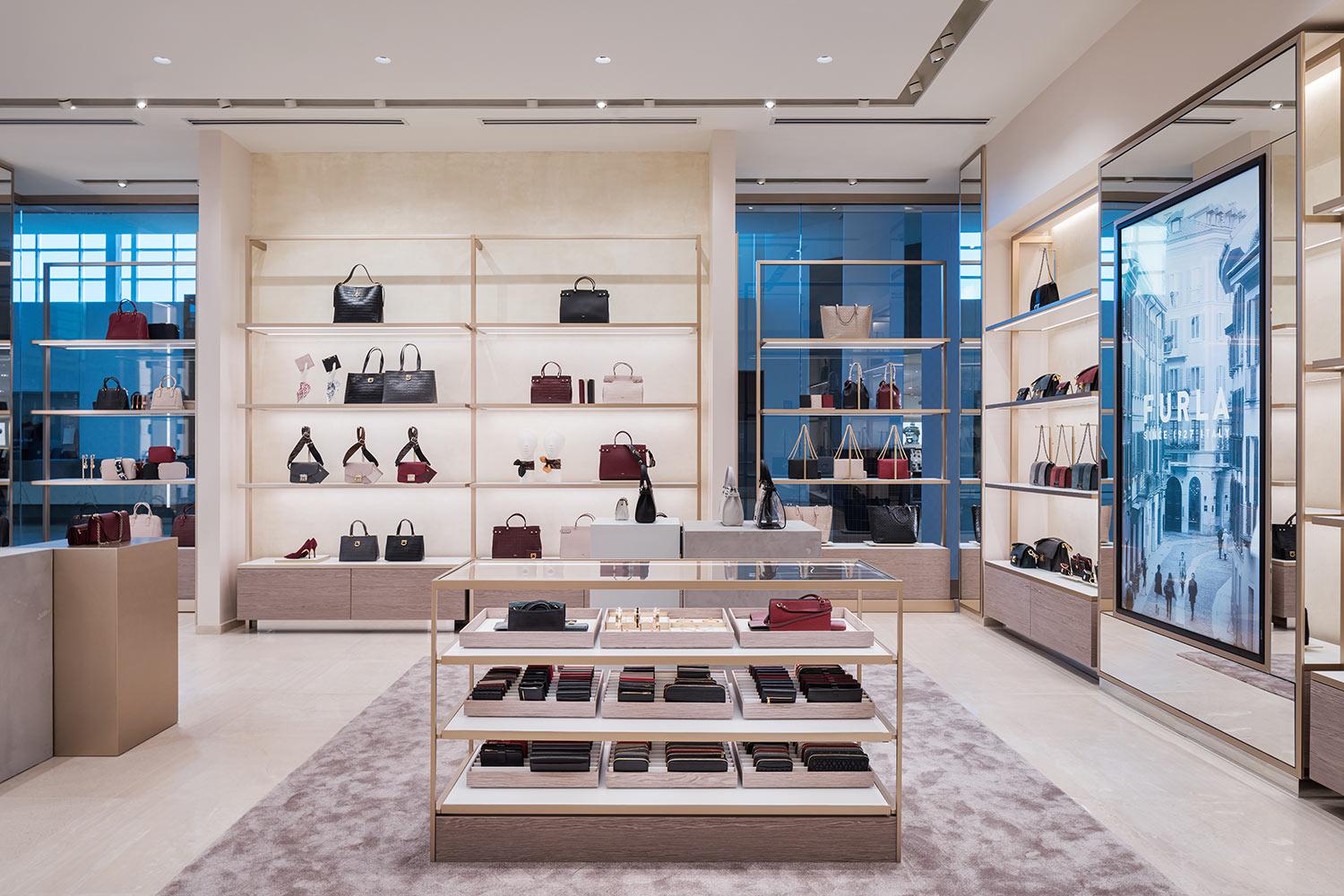 A modern boutique displays handbags, wallets, and accessories on shelves and tables. The store has neutral tones, bright lighting, and a large digital screen showing an advertisement on the right wall.