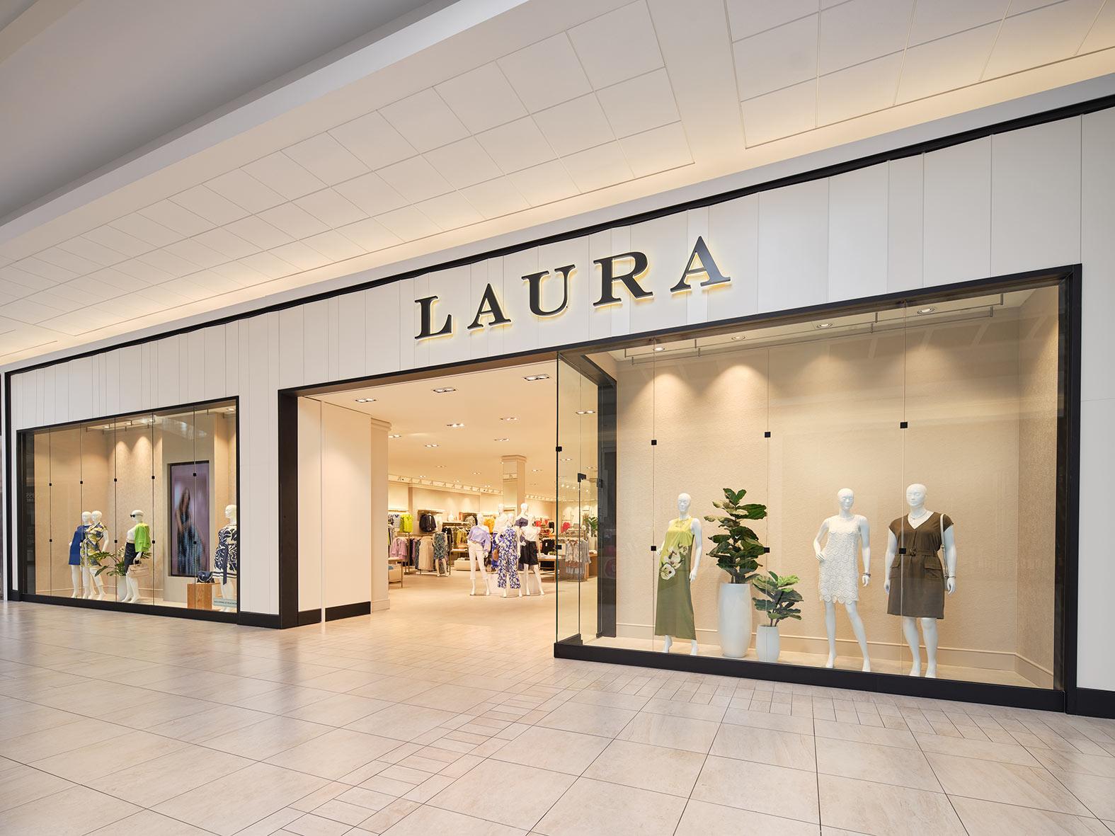 Storefront of a “LAURA” clothing store in a mall, featuring large display windows with mannequins dressed in women’s fashion and a potted plant. The interior displays more clothing and accessories.