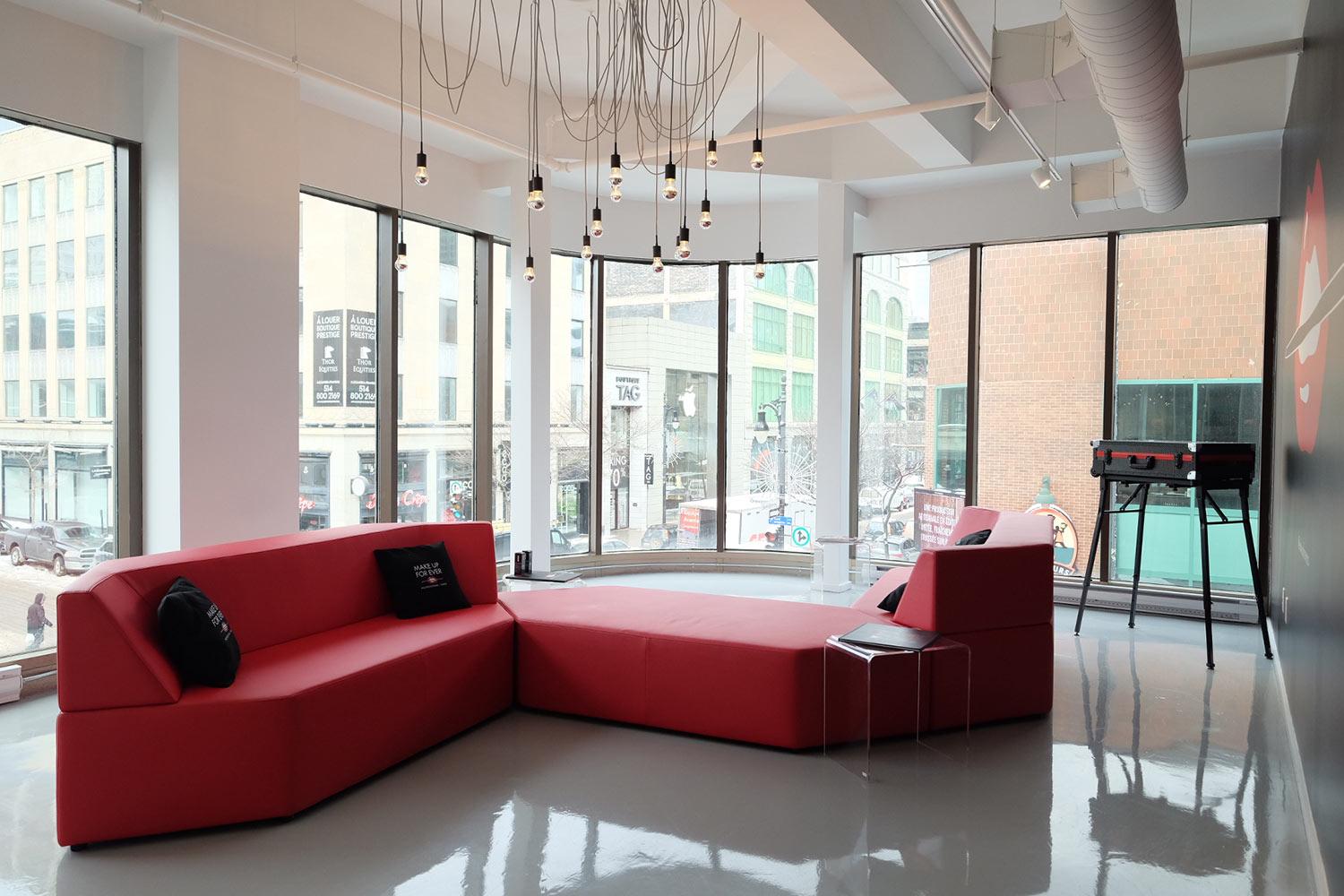 Modern lounge area with large red modular sofas, a small side table, hanging pendant lights, and a foosball table by tall windows overlooking a city street. The floor is shiny and the space is bright and airy.