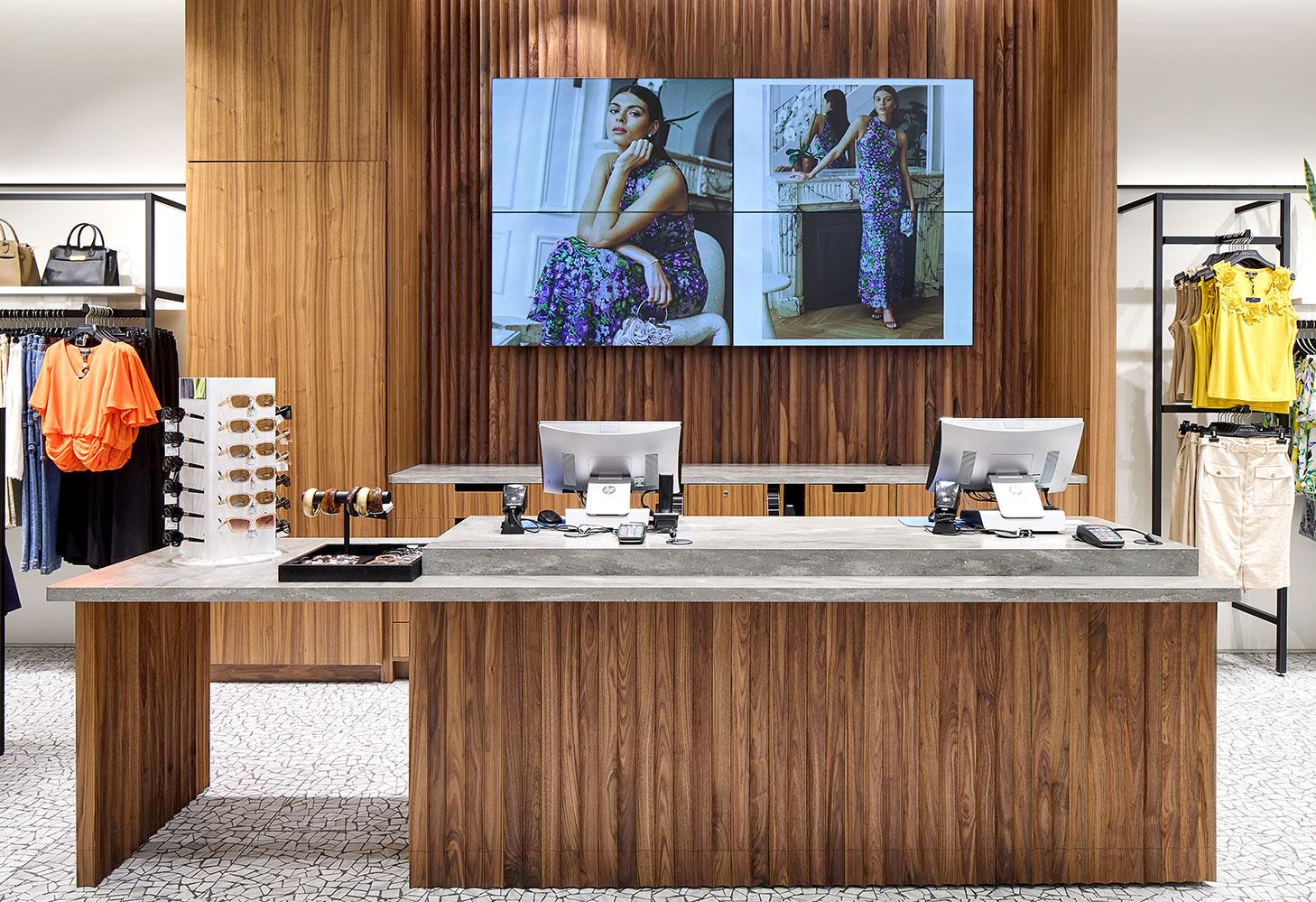 A modern retail store checkout counter with two monitors, a display of sunglasses, and clothing racks. Behind the counter, a large screen shows a woman modeling floral outfits. The decor features wood paneling and a stone countertop.