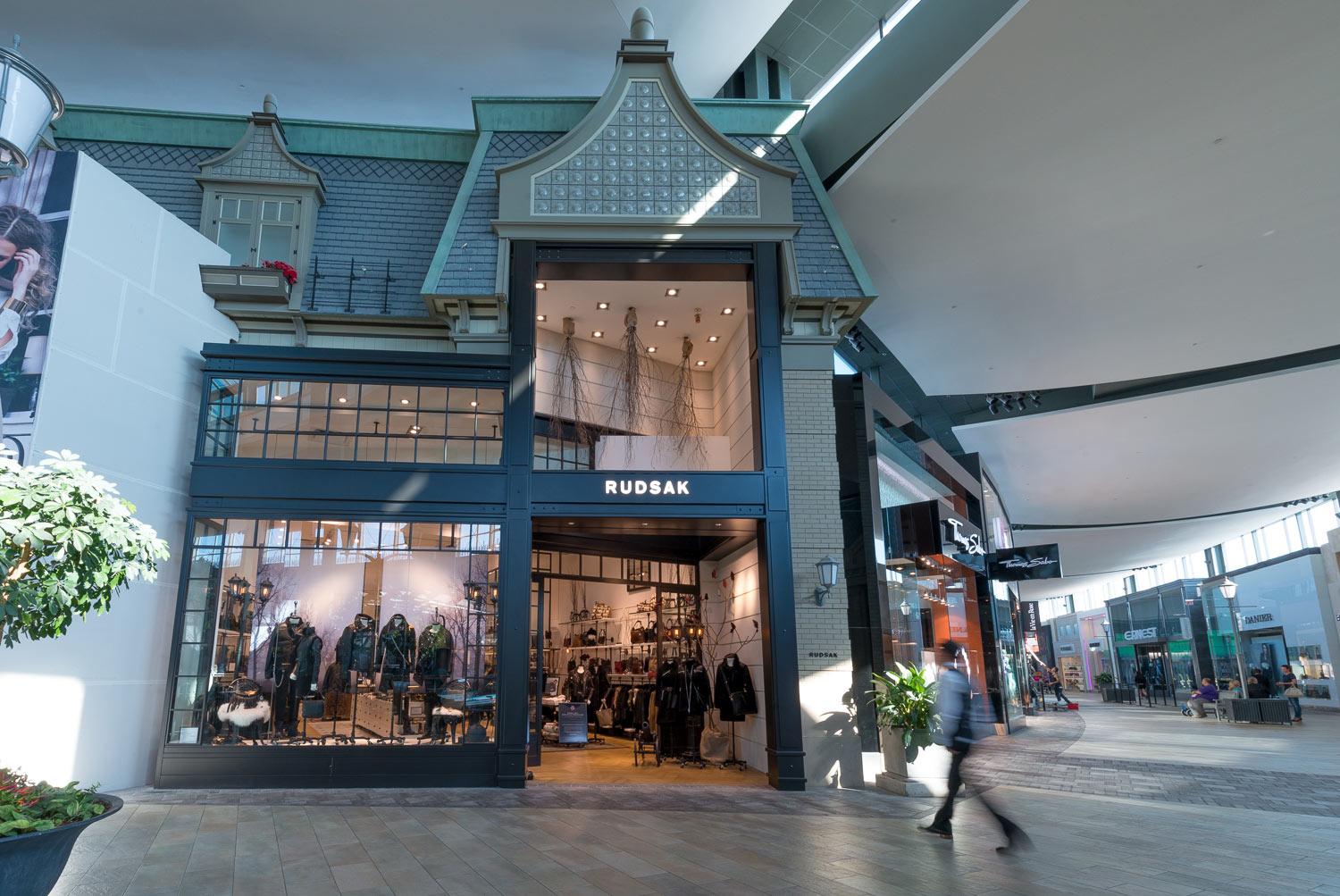 A modern store with large glass windows displays clothing and accessories. The sign above the entrance reads "RUDSAK." A blurred person walks by inside a spacious, well-lit mall.