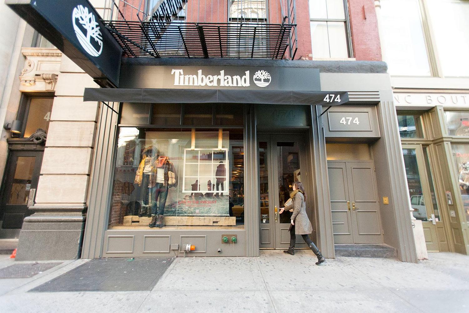A person enters a Timberland store with large display windows featuring mannequins dressed in outdoor clothing and boots. The storefront is gray with the Timberland logo and address number 474 above the door.