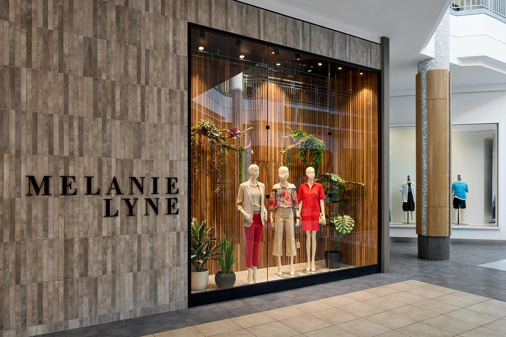 Storefront display of MELANIE LYNE with three mannequins dressed in stylish women's clothing, surrounded by plants and wooden backdrop, inside a modern shopping mall.